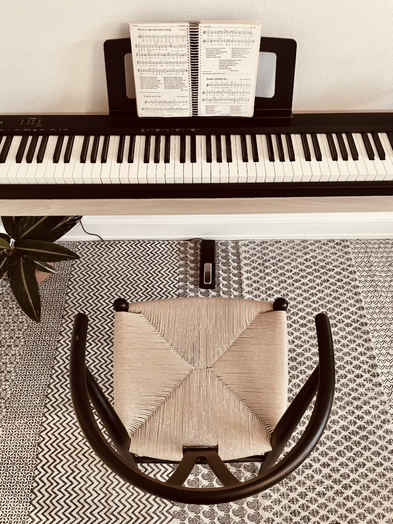 Top view of a wall-mounted digital piano stand in oak with an open songbook and a woven chair below