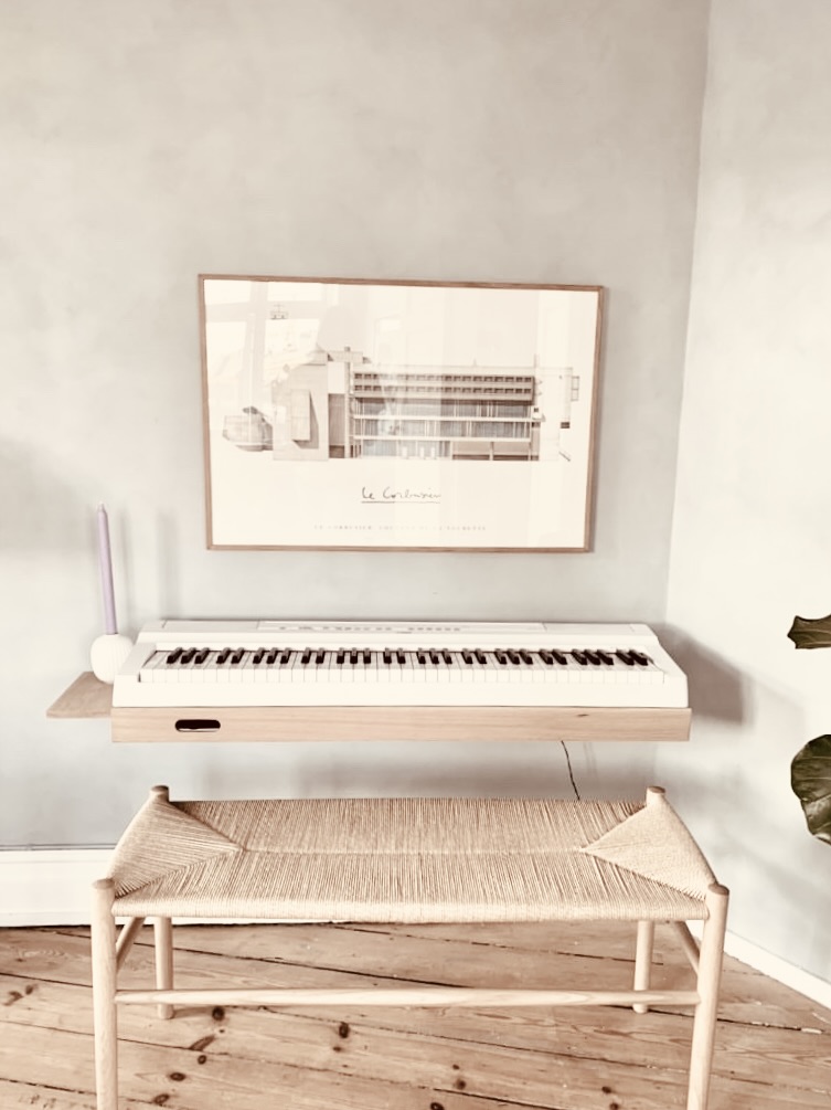 Customer photo of a white digital piano on an oak wall frame below a framed poster with a woven bench