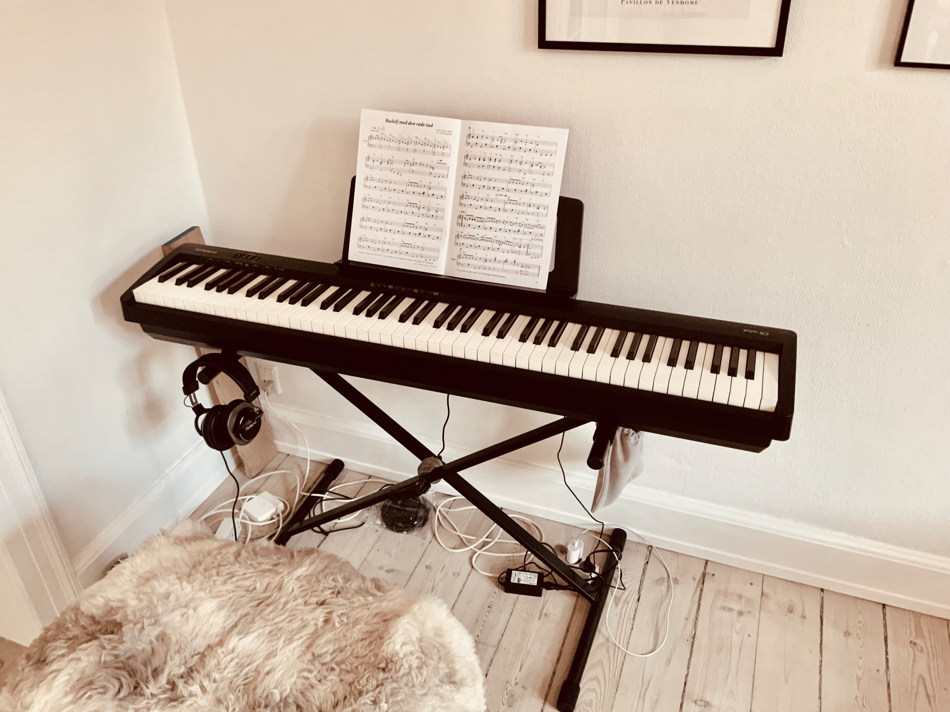 Roland FP-10 digital piano on a black X-stand with sheet music, headphones and cables in a light Scandinavian room