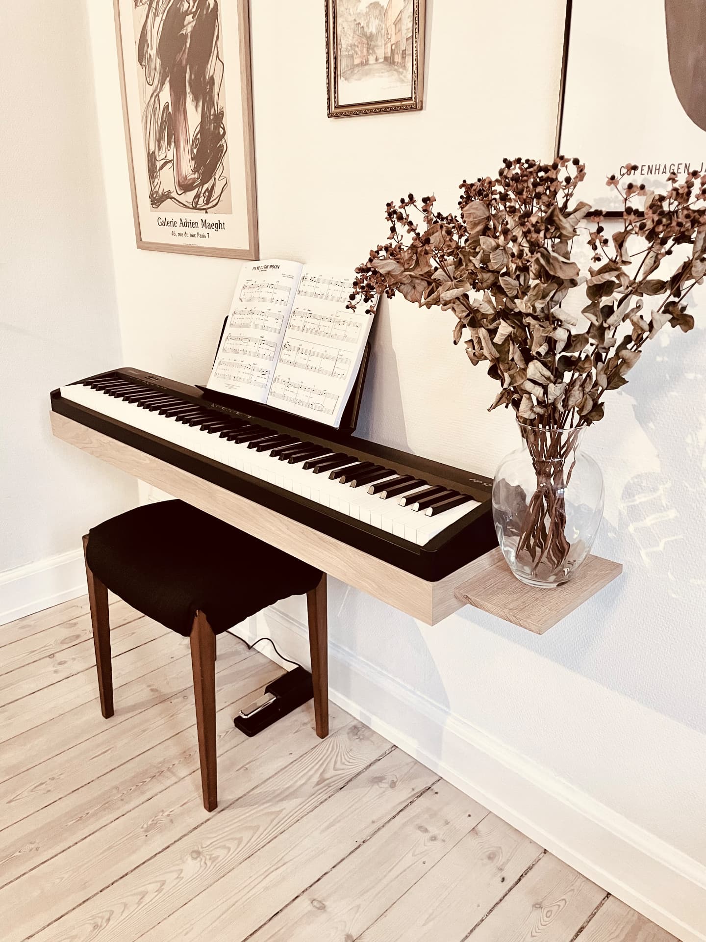 Wall-mounted oak piano stand holding a Roland FP-10 digital piano with side shelf and dried flowers