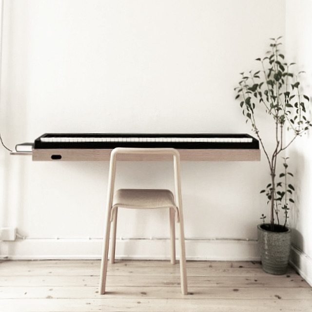 Customer photo of a black digital piano on an oak wall-mounted piano stand in a minimalist white room with a stool and a plant