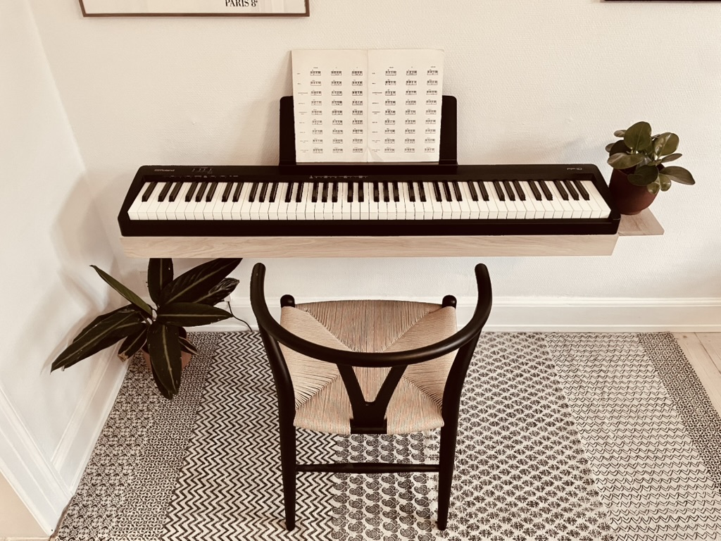 Wall-mounted oak piano stand with a Roland FP-10 digital piano, wishbone chair and plants in a bright Danish living room